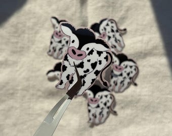 May include: A collection of cartoon cow stickers. Each sticker features a black and white cow with pink accents. The stickers are held by a pair of silver tweezers.