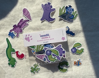 May include: A collection of colorful axolotl stickers, including purple axolotls, a green alligator, and a red shrimp. The stickers are packaged in a clear bag with the text "comfii studio & Axolotl sticker pack 11 pcs".