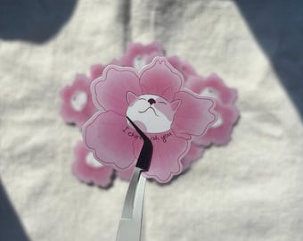 May include: Pink flower-shaped stickers with a cat face in the center and the text "I cherish you". The stickers are being held by tweezers. The stickers are on a white surface.