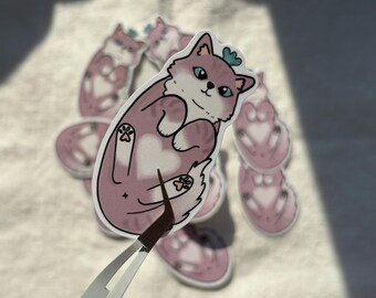 May include: A collection of cat stickers. The stickers feature a pink and white cat with a blue heart-shaped accessory on its head. The cat is lying on its back with its paws visible. The stickers are being held by tweezers.