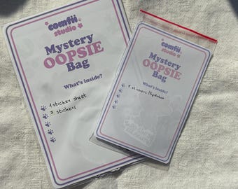 May include: Two cards with the text "comfii studio Mystery OOPSIE Bag" in purple and pink. One card lists "1 sticker sheet" and "7 stickers". The other card lists "8 stickers". The cards are on a white surface.