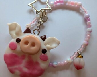 May include: A keychain featuring a sculpted white and pink cow-patterned face with a peach-colored snout, brown horns, and pink cheeks. It is attached to a silver star-shaped clasp and a beaded loop of pink and white beads with a small peach charm.