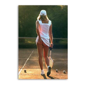 May include: A woman in a white tennis dress and cap, holding a tennis racket, walks on a tennis court. Three tennis balls are on the court. The image is lit by sunlight.
