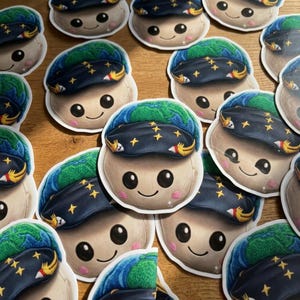 Puede incluir: Una colección de pegatinas con un personaje de dibujos animados sonriente con una cabeza en forma de globo y un sombrero azul oscuro adornado con estrellas doradas. El personaje tiene mejillas rosadas y una expresión alegre. Las pegatinas están sobre una superficie de madera.