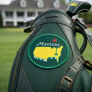 Puede incluir: Una bolsa de golf verde oscuro con el logotipo de Masters. El logotipo presenta un contorno de mapa amarillo y la palabra "Masters" en escritura blanca. Se ven palos de golf, y la bolsa tiene correas de cuero y cremalleras doradas.