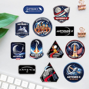 Op de afbeelding: Een verzameling Artemis II-missiestickers met verschillende ontwerpen. De stickers tonen afbeeldingen van de maan, raketten, astronauten en het NASA-logo. Het kleurenpalet omvat blauw, oranje en wit.
