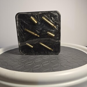 May include: A rectangular black resin block featuring a marbled texture and embedded with six metallic gold-colored bullet casings of varying lengths arranged in a scattered pattern across the front surface of the dark, glossy material.