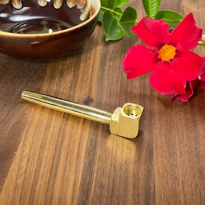 May include: A gold-colored metal pipe with a square bowl and a long, thin stem. The pipe is sitting on a wooden surface.