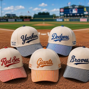 May include: Five baseball caps with off-white crowns and striped brims. The caps feature embroidered team names: Yankees in blue, Dodgers in blue, Red Sox in red, Giants in orange, and Braves in blue, with city names embroidered on the sides.