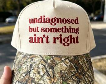 Könnte beinhalten: Eine beige Baseballkappe mit einem Schirm im Camouflage-Muster. Auf der Vorderseite befindet sich der in Rot gestickte Text "undiagnosed but something ain't right" in einer Serifenschrift. Die Kappe hat eine strukturierte Krone und einen Knopf.