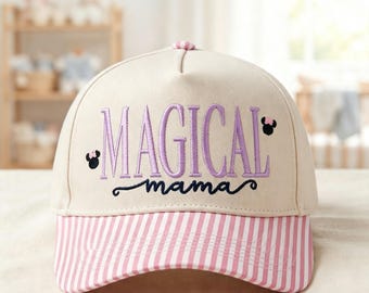 Könnte beinhalten: Eine beige Baseballkappe mit einem rosa-weiß gestreiften Schirm. Auf der Vorderseite befindet sich der gestickte Schriftzug "MAGICAL" in Lila und "mama" in Dunkelblau, akzentuiert durch zwei kleine schwarze Mausohren mit rosa Schleifen.