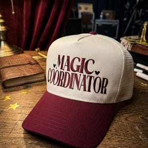 May include: A cream-colored trucker hat with a burgundy brim and the words "MAGIC COORDINATOR" embroidered in burgundy script. The hat has a mesh back and a small burgundy button on top. The text is flanked by small black hearts.