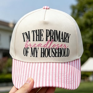 May include: A cream-colored baseball cap with a pink and white striped brim. The front features black embroidered text that reads "I'M THE PRIMARY" and "OF MY HOUSEHOLD," with the word "breadloser" embroidered in pink script between the lines.
