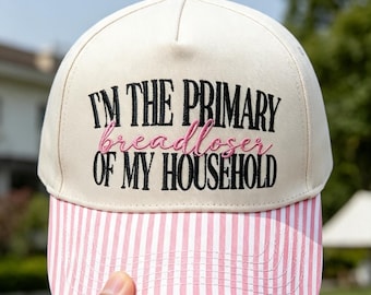 Könnte beinhalten: Eine cremefarbene Baseballkappe mit einem rosa-weiß gestreiften Schirm. Auf der Vorderseite befindet sich ein schwarzer Sticktext mit der Aufschrift "I'M THE PRIMARY" und "OF MY HOUSEHOLD", wobei das Wort "breadloser" in rosa Schreibschrift dazwischen liegt.