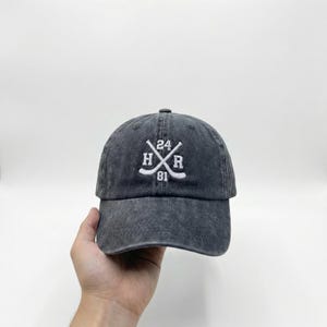 May include: A dark gray baseball cap with a curved brim. The cap features white embroidered text and graphics, including the letters "H" and "R", crossed hockey sticks, and the numbers "24" and "81".