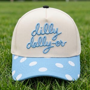 May include: A cream-colored baseball cap with a black button on top. The front features the text "dilly dally-er" embroidered in light blue script. The brim is light blue with a white polka dot pattern across the entire surface.