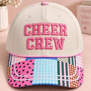 K&ouml;nnte beinhalten: Eine cremefarbene Baseballkappe mit dem in rosa Blockbuchstaben gestickten Text CHEER CREW auf der Vorderseite. Der Schirm zeigt ein buntes Patchwork-Muster aus rosa Punkten, blauem Vichy-Karo, gr&uuml;nen Streifen und braunem Karomuster.