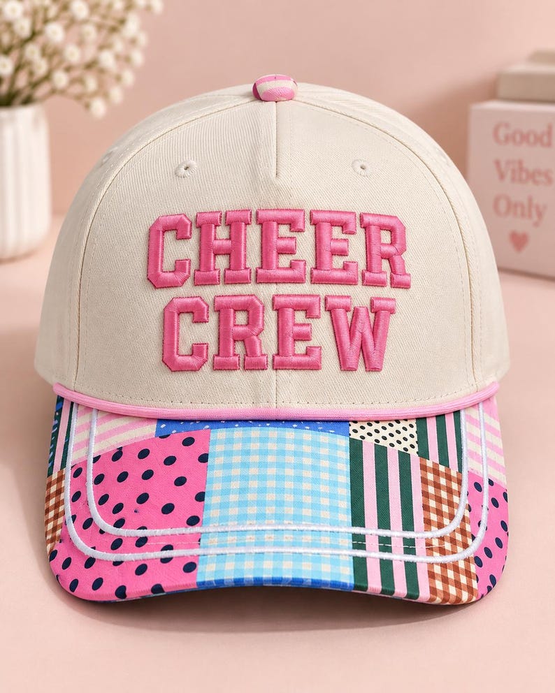 K&ouml;nnte beinhalten: Eine cremefarbene Baseballkappe mit dem in rosa Blockbuchstaben gestickten Text CHEER CREW auf der Vorderseite. Der Schirm zeigt ein buntes Patchwork-Muster aus rosa Punkten, blauem Vichy-Karo, gr&uuml;nen Streifen und braunem Karomuster.