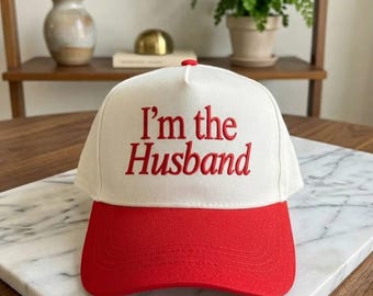 Könnte beinhalten: Eine weiße Baseballkappe mit rotem Schirm und rotem gesticktem Text auf der Vorderseite, der I'm the Husband lautet. Die Kappe verfügt über ein strukturiertes Vorderteil und einen Knopf oben in der Mitte in einem zweifarbigen Design.
