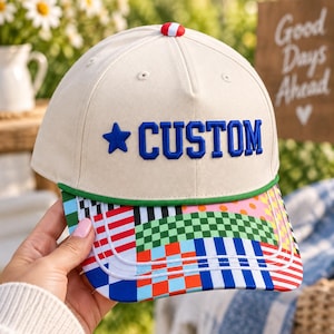 K&ouml;nnte beinhalten: Eine cremefarbene Baseballkappe mit dem Wort CUSTOM in blauen Stickbuchstaben neben einem blauen Stern. Die Kappe hat einen gr&uuml;nen Rand und einen Schirm, der mit einem bunten Patchwork-Muster aus Karos, Streifen und Punkten verziert ist.