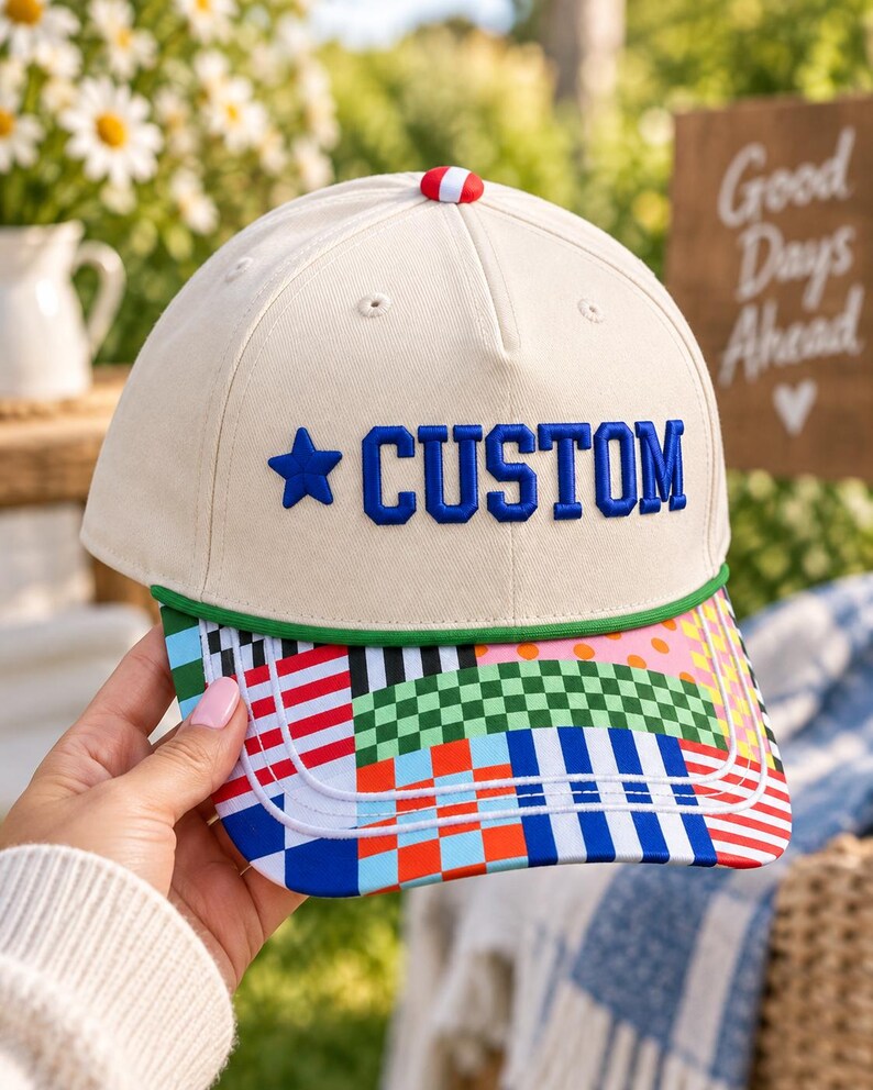 K&ouml;nnte beinhalten: Eine cremefarbene Baseballkappe mit dem Wort CUSTOM in blauen Stickbuchstaben neben einem blauen Stern. Die Kappe hat einen gr&uuml;nen Rand und einen Schirm, der mit einem bunten Patchwork-Muster aus Karos, Streifen und Punkten verziert ist.
