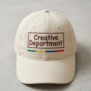 May include: A cream-colored baseball cap with the words "Creative Department" embroidered in brown, enclosed in a brown rectangle. Below the text is a horizontal stripe of green, yellow, blue, and red. The cap has a curved brim.