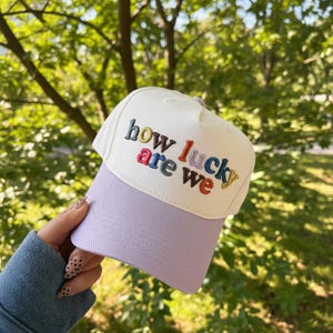 May include: A white baseball cap with a lavender brim. The cap features the embroidered phrase "how lucky are we" in colorful letters. The background is a blurred view of green foliage.