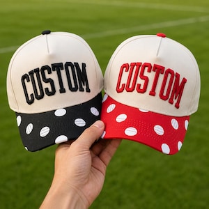 May include: Two baseball caps with off-white crowns and the word CUSTOM embroidered on the front. One cap features a black brim with white polka dots, while the other has a red brim with white polka dots. Both caps have a button on top.
