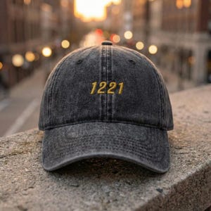 May include: A dark gray baseball cap with the embroidered number "1221" in gold thread. The cap has a curved brim and a slightly faded, vintage look. The background is blurred, suggesting an outdoor setting.