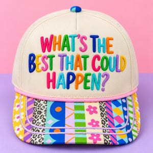 May include: A cream-colored baseball cap featuring the embroidered text "WHAT'S THE BEST THAT COULD HAPPEN?" in multicolored letters. The brim is decorated with a vibrant patchwork pattern of stripes, leopard print, and flowers.