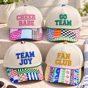 May include: Four off-white baseball caps with embroidered text and colorful patterned brims. The caps feature the phrases CHEER BABE, GO TEAM, TEAM JOY, and FAN CLUB in pink, green, blue, and yellow lettering with matching top buttons.