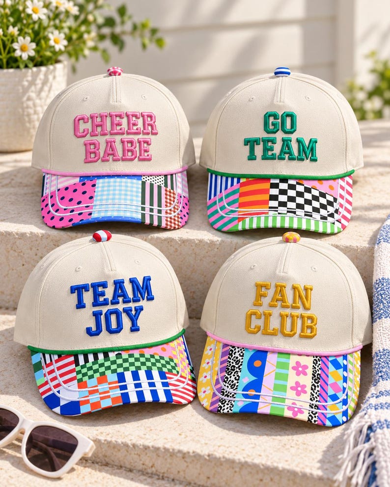 K&ouml;nnte beinhalten: Vier cremefarbene Baseballkappen mit gesticktem Text und bunten gemusterten Schirmen. Die Kappen tragen die Aufschriften CHEER BABE, GO TEAM, TEAM JOY und FAN CLUB in rosa, gr&uuml;ner, blauer und gelber Schrift mit passenden Kn&ouml;pfen oben.