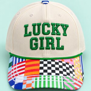 K&ouml;nnte beinhalten: Eine cremefarbene Baseballkappe mit dem in gr&uuml;nen Buchstaben gestickten Text LUCKY GIRL. Die Kappe hat eine gr&uuml;ne Kordel &uuml;ber dem Schirm, einen blau-wei&szlig; gestreiften Knopf oben und einen mehrfarbigen Schirm mit Karomustern und Streifen.