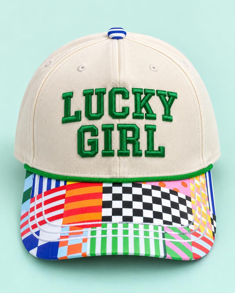 K&ouml;nnte beinhalten: Eine cremefarbene Baseballkappe mit dem in gr&uuml;nen Buchstaben gestickten Text LUCKY GIRL. Die Kappe hat eine gr&uuml;ne Kordel &uuml;ber dem Schirm, einen blau-wei&szlig; gestreiften Knopf oben und einen mehrfarbigen Schirm mit Karomustern und Streifen.