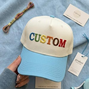 以下が含まれることがあります： ライトブルーのつばとクリーム色のクラウンが付いた野球帽。「CUSTOM」の文字が虹色で前面に刺繍されています。カラフルな刺繍糸の束と製品タグも表示されています。