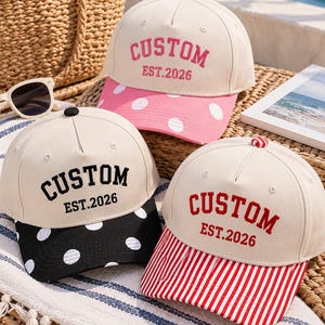 May include: Three baseball caps with cream-colored crowns and embroidered text reading CUSTOM EST.2026. One cap has a pink polka-dot brim, one has a black polka-dot brim, and one has a red and white striped brim. Each cap features a small button on top.