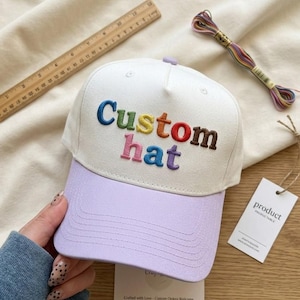 以下が含まれることがあります： 虹色の「Custom hat」の刺繍が入った、ライラックのつばが付いた白い野球帽。帽子は人が持っています。木製の定規と刺繍糸が背景にあります。