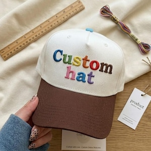 May include: A white and brown baseball cap with the embroidered text "Custom hat" in rainbow colors. The cap has a brown brim and a light blue button on top. A wooden ruler and a spool of colorful thread are in the background.