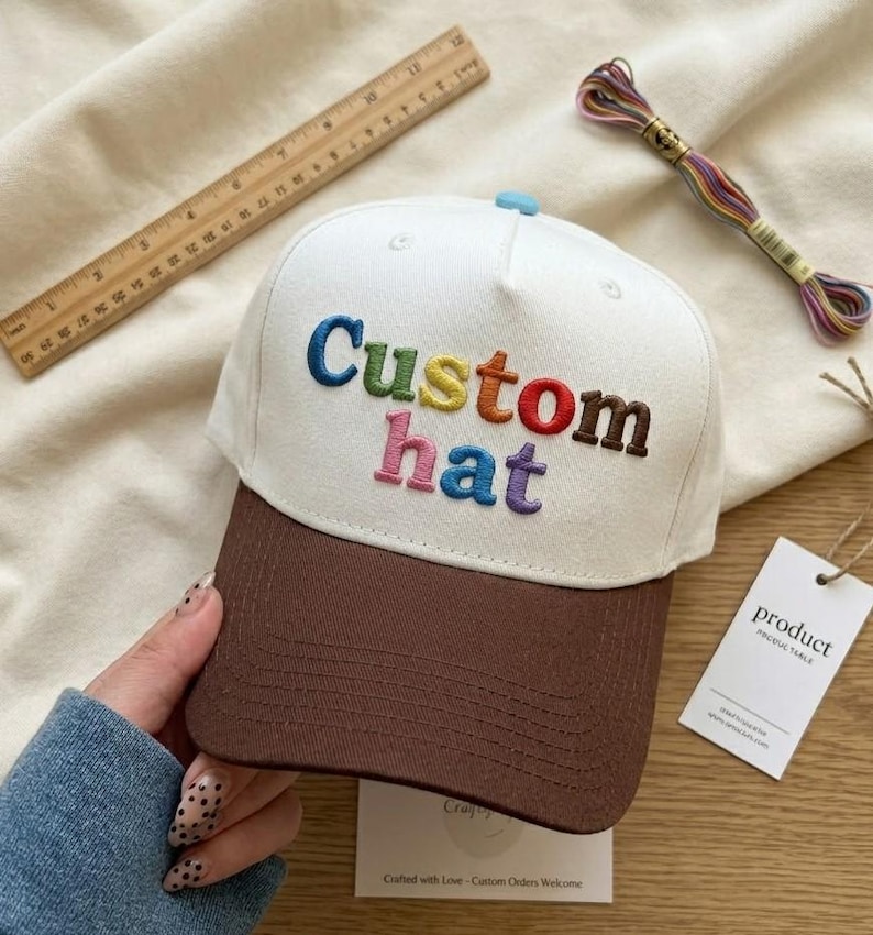 以下が含まれることがあります： 虹色の「Custom hat」の刺繍が入った白と茶色の野球帽。 つばは茶色で、上部に水色のボタンが付いています。 木製の定規とカラフルな糸巻きが背景にあります。