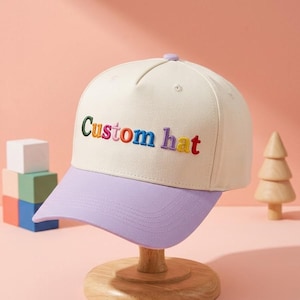 以下が含まれることがあります： クリーム色の野球帽、ラベンダー色のつば付き。カラフルな文字で「Custom hat」と刺繍されています。ピンク色の背景に、木製のスタンドに帽子が飾られています。