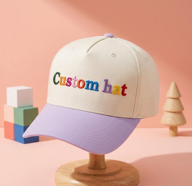 以下が含まれることがあります： クリーム色の野球帽、ラベンダー色のつば付き。カラフルな文字で「Custom hat」と刺繍されています。ピンク色の背景に、木製のスタンドに帽子が飾られています。