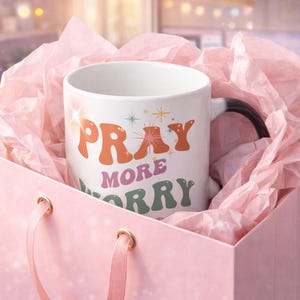May include: A white ceramic mug with the words "PRAY MORE WORRY LESS" in orange, pink, and green lettering. The mug has a black handle and is inside a pink gift bag with pink tissue paper.