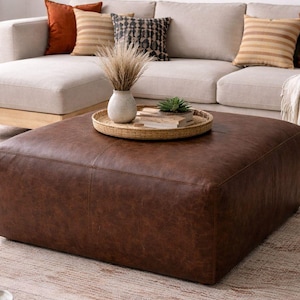 May include: A large, square, brown leather ottoman sits in front of a beige sectional sofa. Decorative cushions in shades of orange, beige, and black adorn the sofa. A woven tray with a vase and small plant rests on the ottoman. The room has a neutral colour palette.