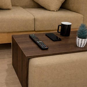 May include: A contemporary living room setup with a beige sofa and cushions. A dark brown coffee table displays a remote control, smartphone, black mug, and a small potted plant. The table rests on a matching ottoman, creating a coordinated and stylish environment.