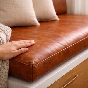 May include: A brown leather bench cushion with a hand resting on it. The cushion is rectangular and has a stitched border. Two beige pillows are in the background. The bench is part of a window seat with wooden drawers.