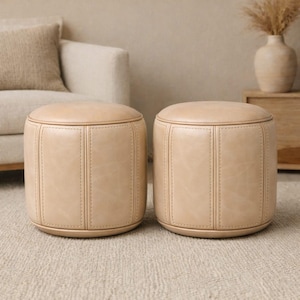 May include: Two beige, cylindrical ottomans with stitched detailing. The ottomans have a rounded top and base, and are placed on a neutral-colored rug. The background includes a beige sofa, a wooden side table, and decorative vase.