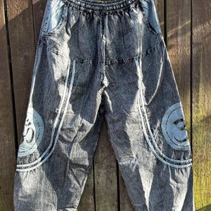 Può includere: Pantaloni harem grigi con un aspetto sbiadito e strutturato. I pantaloni presentano dettagli azzurri, tra cui disegni circolari con un simbolo e strisce verticali. Appesi a una gruccia bianca su uno sfondo di legno.