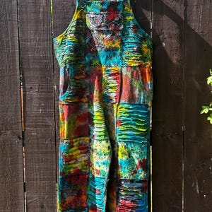 May include: A patchwork jumpsuit in a vibrant array of colours, including red, blue, green, and yellow. The garment features a bib-style top with adjustable straps and pockets. It is hanging on a black hanger against a wooden fence, with a mix of patterns.