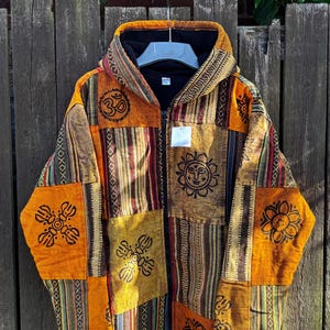 May include: A hooded patchwork jacket in shades of orange, brown, and beige, with a zip closure. The jacket is decorated with various patterns, stripes, and symbols, including a sun face and the Om symbol. The jacket is hanging on a wooden fence.