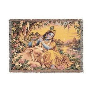 May include: A rectangular woven tapestry blanket featuring a blue-skinned figure playing a golden flute while seated beside a white cow. The scene includes lush foliage, pink flowers, a calm river, and a distant temple under a warm golden sky.